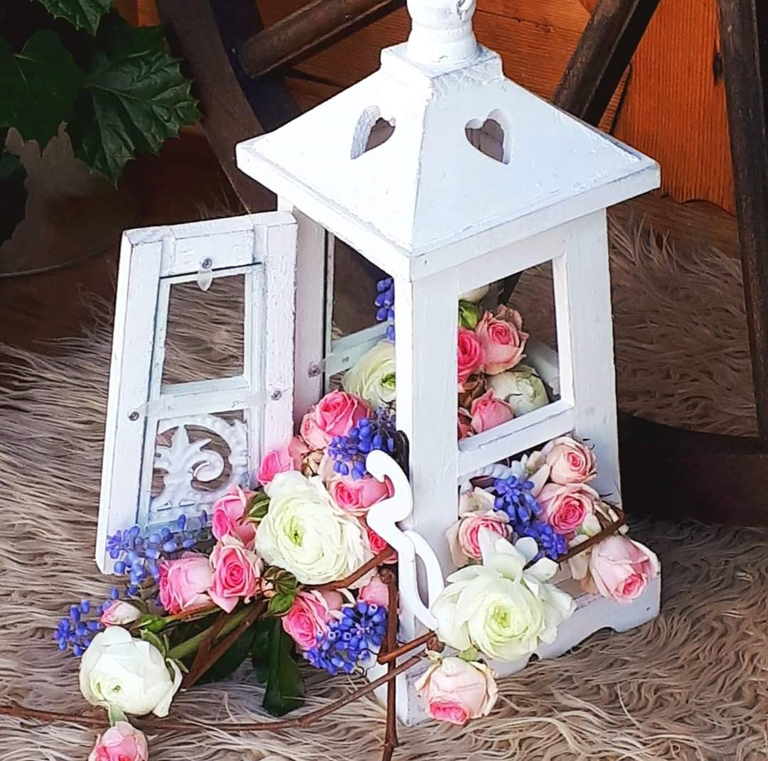 Biała, drewniana latarenka z otwartymi drzwiczkami, wypełniona różowymi i białymi różami oraz niebieskimi hiacyntami na futrzanym tle, z dekoracyjnym, białym łabędziem na zewnątrz.