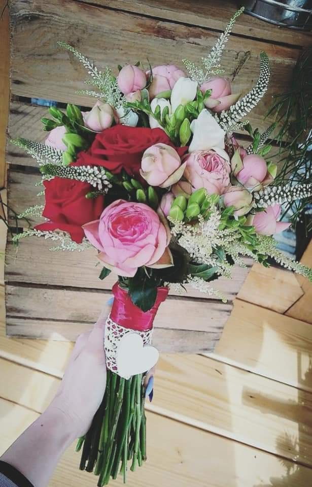 Ręcznie trzymany bukiet z różowych i czerwonych róż, dekorowany białą koronką i sercem na tle drewnianych desek.