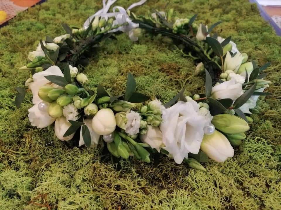Wieniec z białych kwiatów (głównie frezji i tulipanów) z zielonymi liśćmi i perłami, ułożony na tle z mchu.