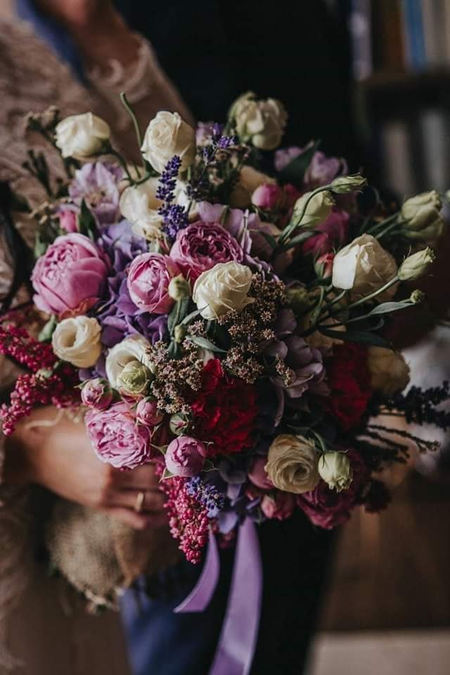 Delikatny bukiet mieszany z różowych piwonii, kremowych róż, fioletowych eustom i lawendy, przewiązany satynową wstążką w odcieniach fioletu, trzymany w dłoniach z widocznym złotym pierścionkiem.