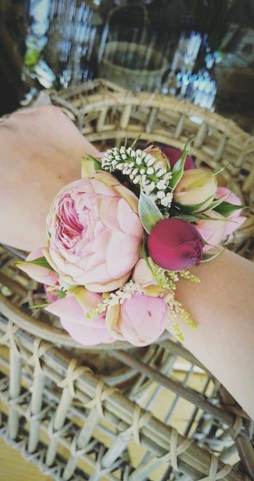 Kwiatowa bransoletka na nadgarstku, z bladoróżową piwonią, bordowym pąkiem róży i białymi drobnymi kwiatkami, na tle wiklinowego kosza.