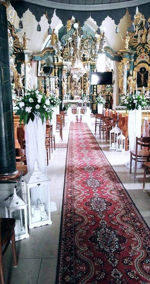 Uroczysta dekoracja kościoła z czerwonym dywanem, białymi lampionami i kompozycjami kwiatowymi przy ławkach.