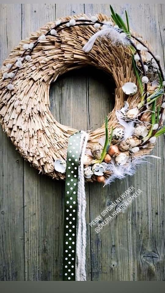Słomiany wieniec wielkanocny z jajkami przepiórczymi, piórkami i gałązkami wierzby, zawieszony na drewnianym tle, z zieloną wstążką w białe kropki.