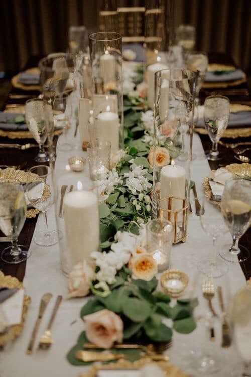 Uroczysty stół z białym obrusem, świecami w szklanych cylindrach i złotych lampionach, bukietami z białych i brzoskwiniowych róż oraz zielonymi liśćmi, nakryty złotymi talerzami i kieliszkami.