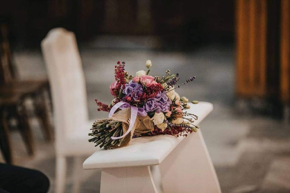 Bukiet ślubny z fioletowymi i różowymi kwiatami, przewiązany jutowym materiałem i fioletową wstążką, leży na białym krześle w kościele.