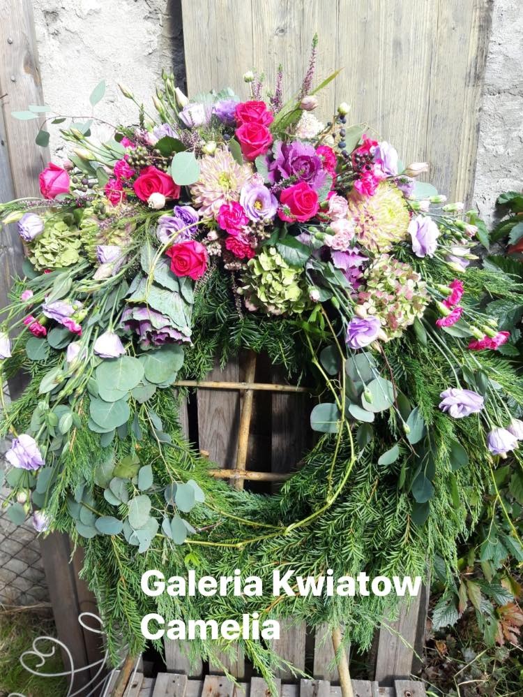 Wieniec z gałązek świerkowych i eukaliptusa, udekorowany różowymi różami, fioletowymi eustomami i zielonymi hortensjami, oparty o drewniane drzwi, z logo 'Galeria Kwiatów Camelia' w dolnej części.