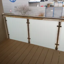 Balkon z podłogą z desek kompozytowych i balustradą ze stali nierdzewnej z wypełnieniem z matowego szkła, widok na plac budowy.