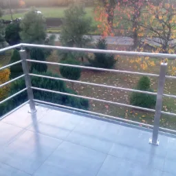Balkon z balustradą nierdzewną, widok na ogród z drzewami o jesiennych liściach, płytki podłogowe w odcieniach szarości.