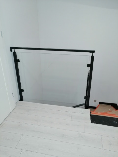 Szklana balustrada z czarnymi elementami mocującymi zamontowana na schodach w trakcie wykańczania, widoczny fragment podestu z zabezpieczeniem krawędzi.