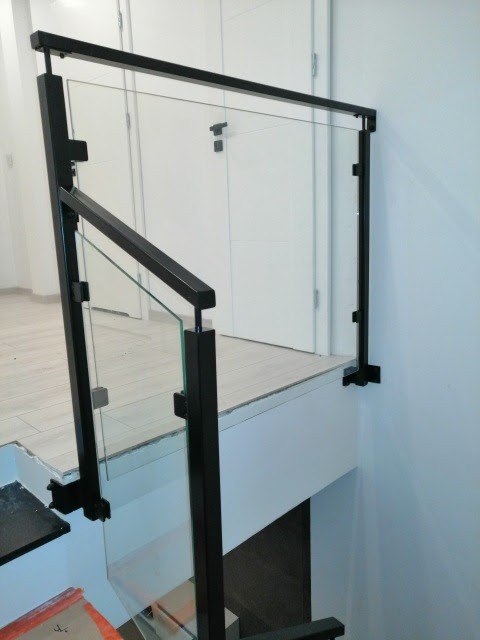 Szklana balustrada schodowa z czarnymi, metalowymi słupkami i poręczą, widok na białe drzwi i jasne podłogi.