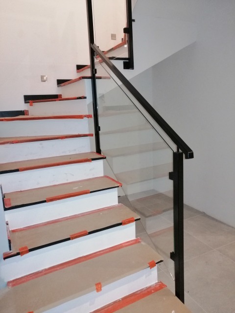 Szklana balustrada z czarnymi elementami mocującymi na schodach z zabezpieczeniami na stopniach podczas montażu.