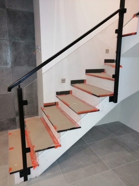 Nowoczesne schody z czarnymi metalowymi elementami i szklaną balustradą, zabezpieczone folią ochronną na stopniach, widoczne szare płytki podłogowe.