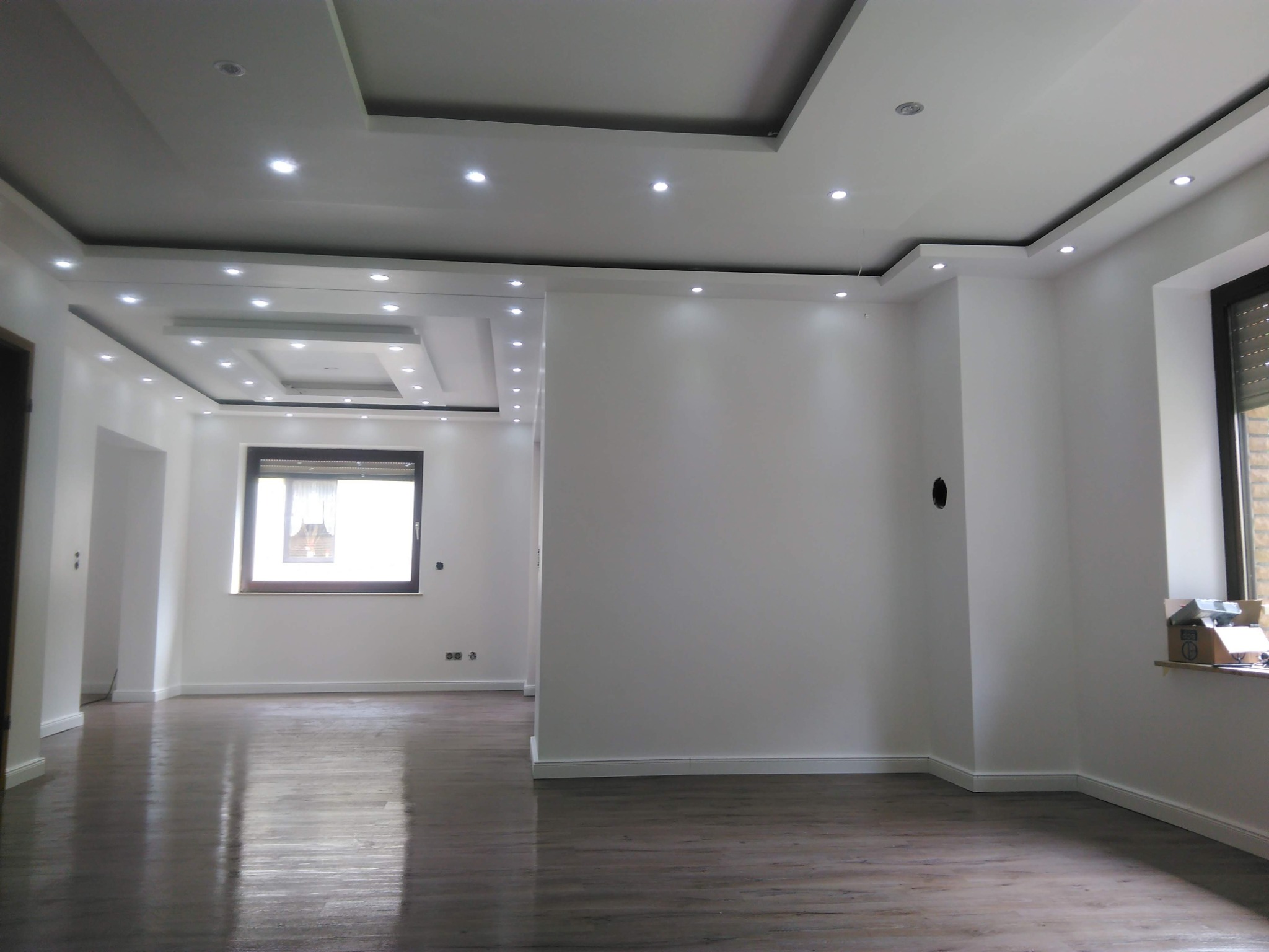 Jasne, przestronne wnętrze po remoncie z drewnopodobną podłogą, białymi ścianami, oknami z roletami i sufitem z wbudowanym oświetleniem punktowym.