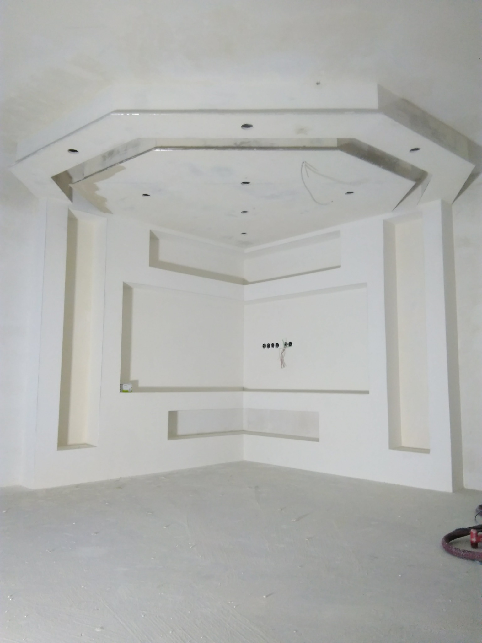 Wnętrze pokoju w trakcie wykończenia, z widoczną konstrukcją z płyt gipsowo-kartonowych na suficie i ścianach, przygotowaną pod montaż oświetlenia i sprzętu RTV. Na podłodze widoczne ślady po...