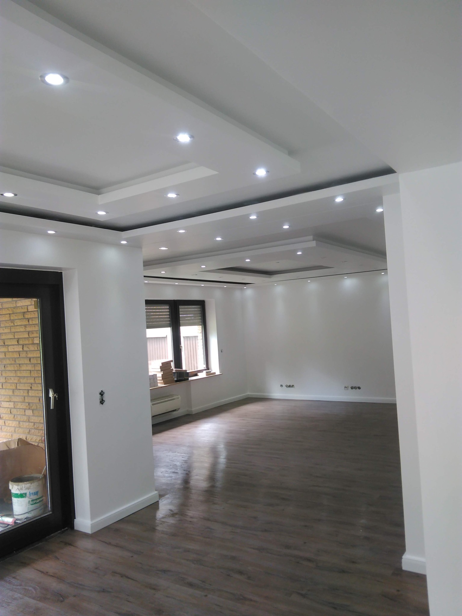 Jasne, przestronne wnętrze po remoncie: parkiet w odcieniach brązu, białe ściany z dekoracyjnym oświetleniem sufitowym i listwami, okno z żaluzjami i grzejnikiem pod parapetem, widoczna puszka...