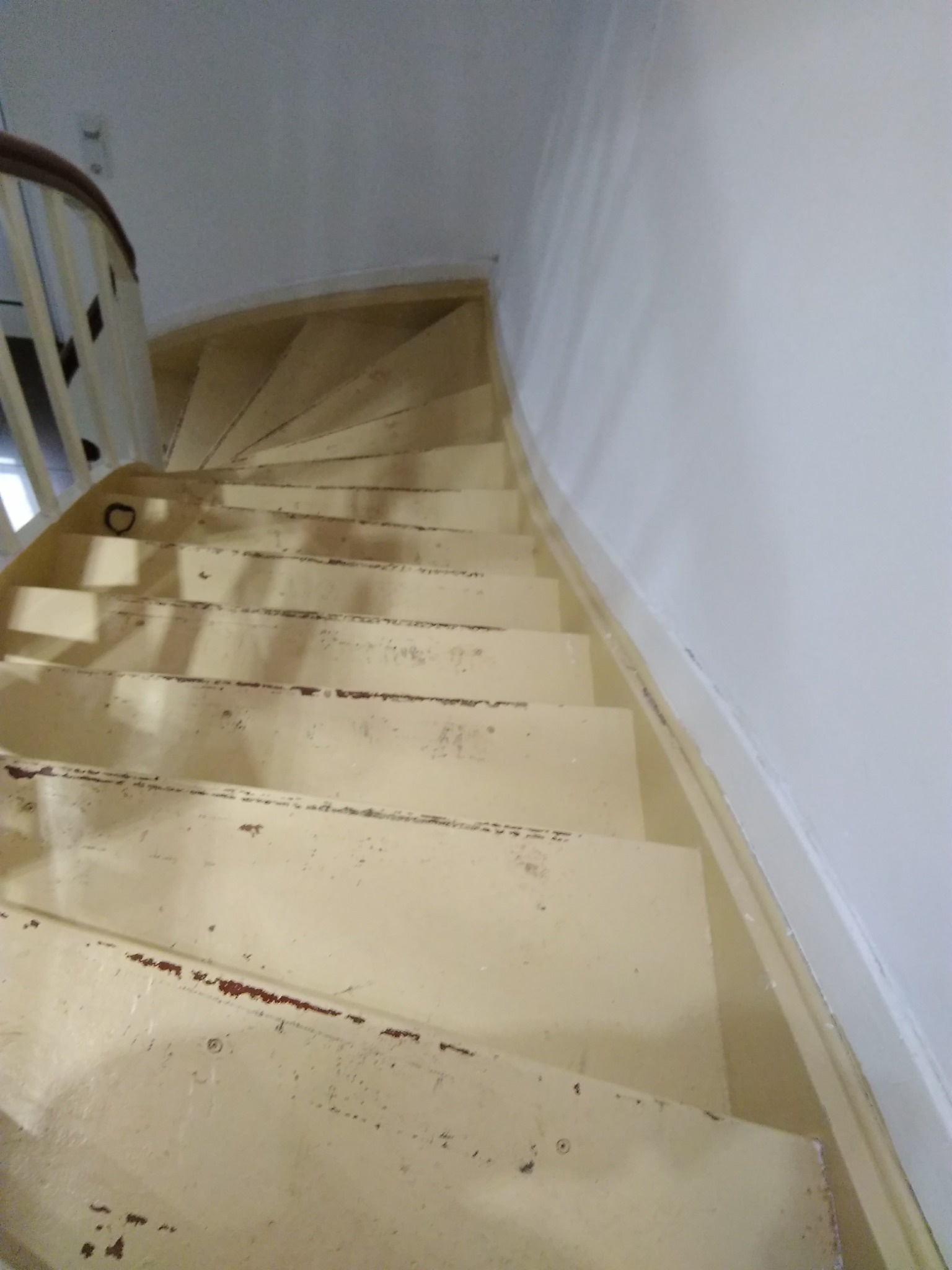 Widok z góry na zakrzywione, drewniane schody z odpryskami farby, prowadzące w górę wzdłuż białej ściany, z widoczną drewnianą balustradą po lewej stronie.