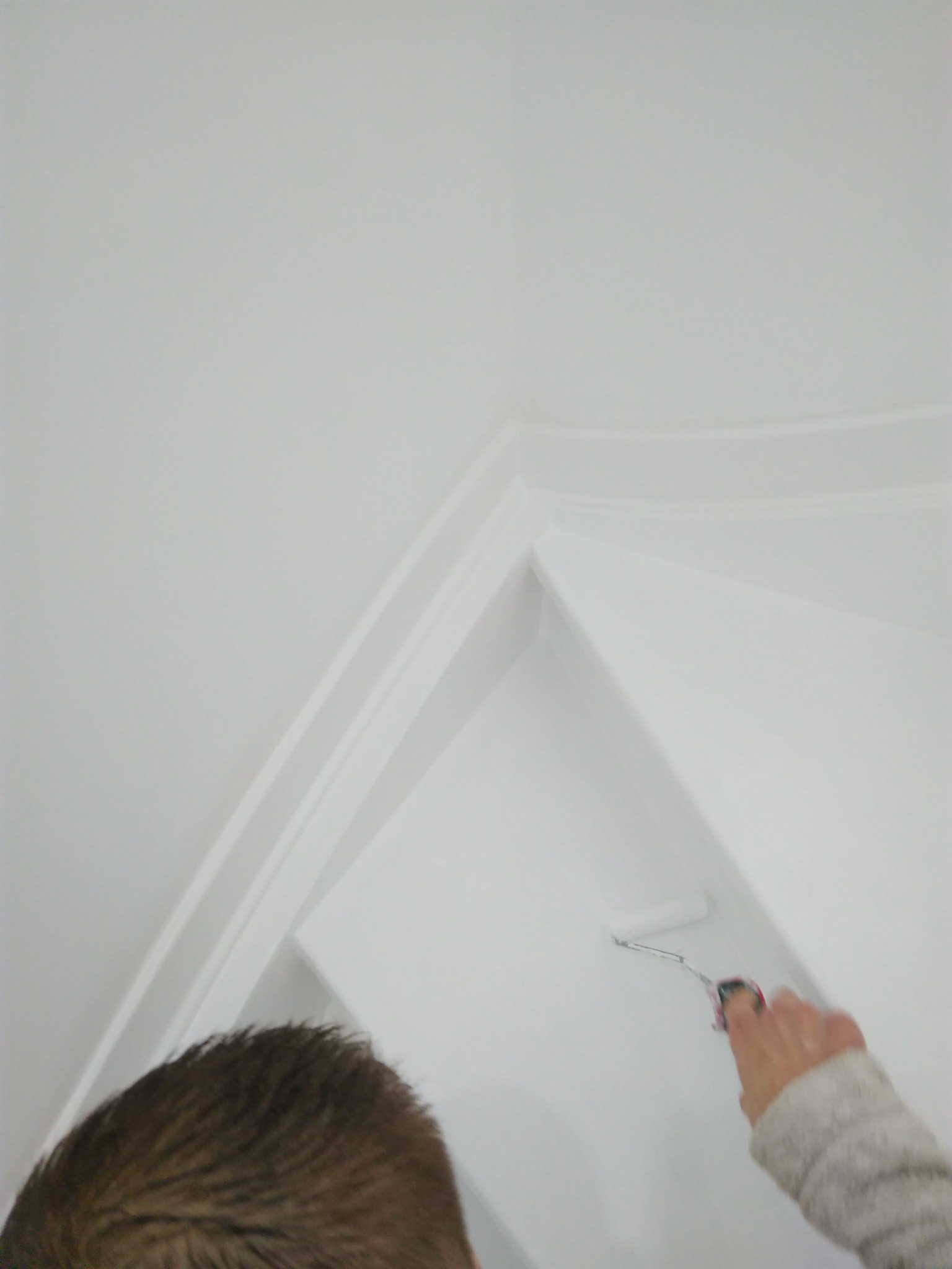 Malowanie białej ściany i sufitu wałkiem z widocznym fragmentem głowy malarza i ręką w swetrze, kąt ujęcia od dołu.