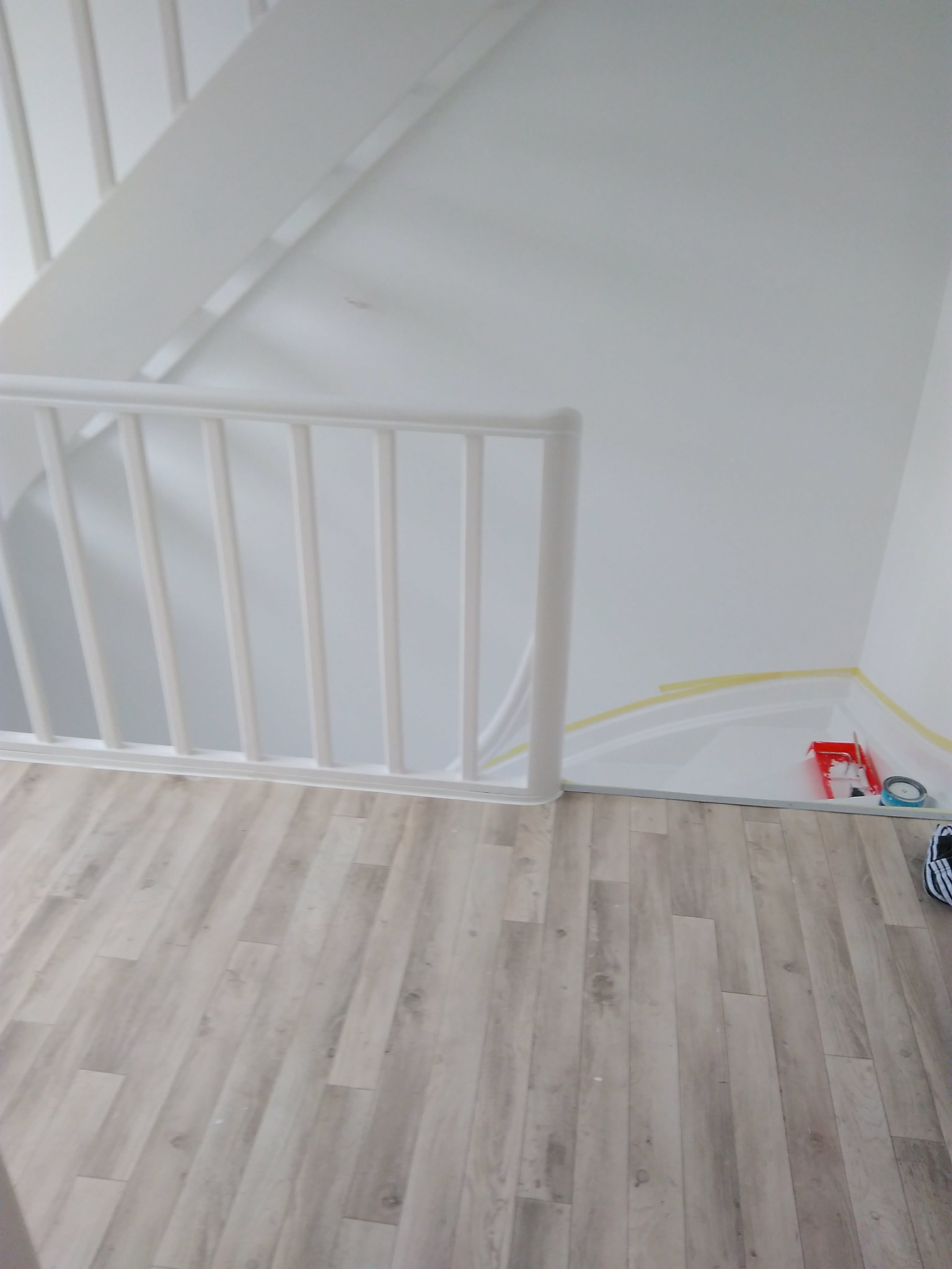 Białe schody z drewnianą podłogą, ściany zabezpieczone taśmą malarską, widoczne akcesoria malarskie.