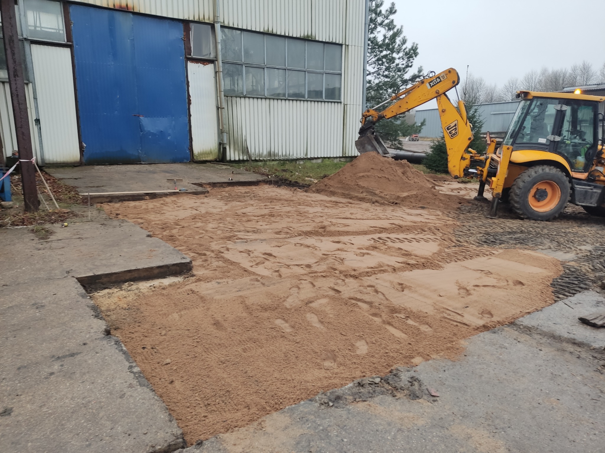 Żółta koparko-ładowarka JCB przygotowuje podłoże z piasku pod przyszłą kostkę brukową przed budynkiem z niebieską bramą.