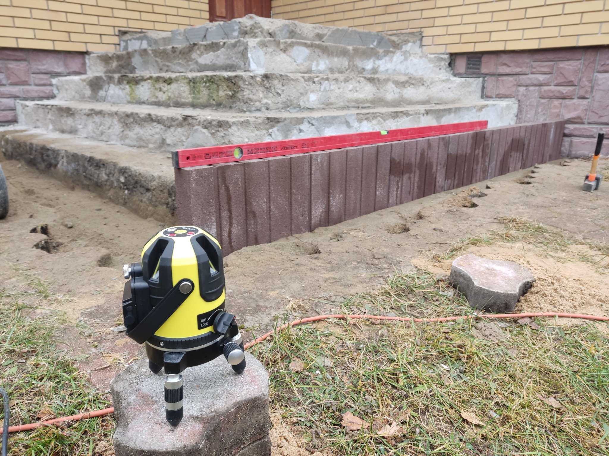 Układanie obrzeży z kostki brukowej w Hajnówce, widoczny laser krzyżowy na kamieniu i poziomica na obrzeżu, w tle schody wejściowe do domu.