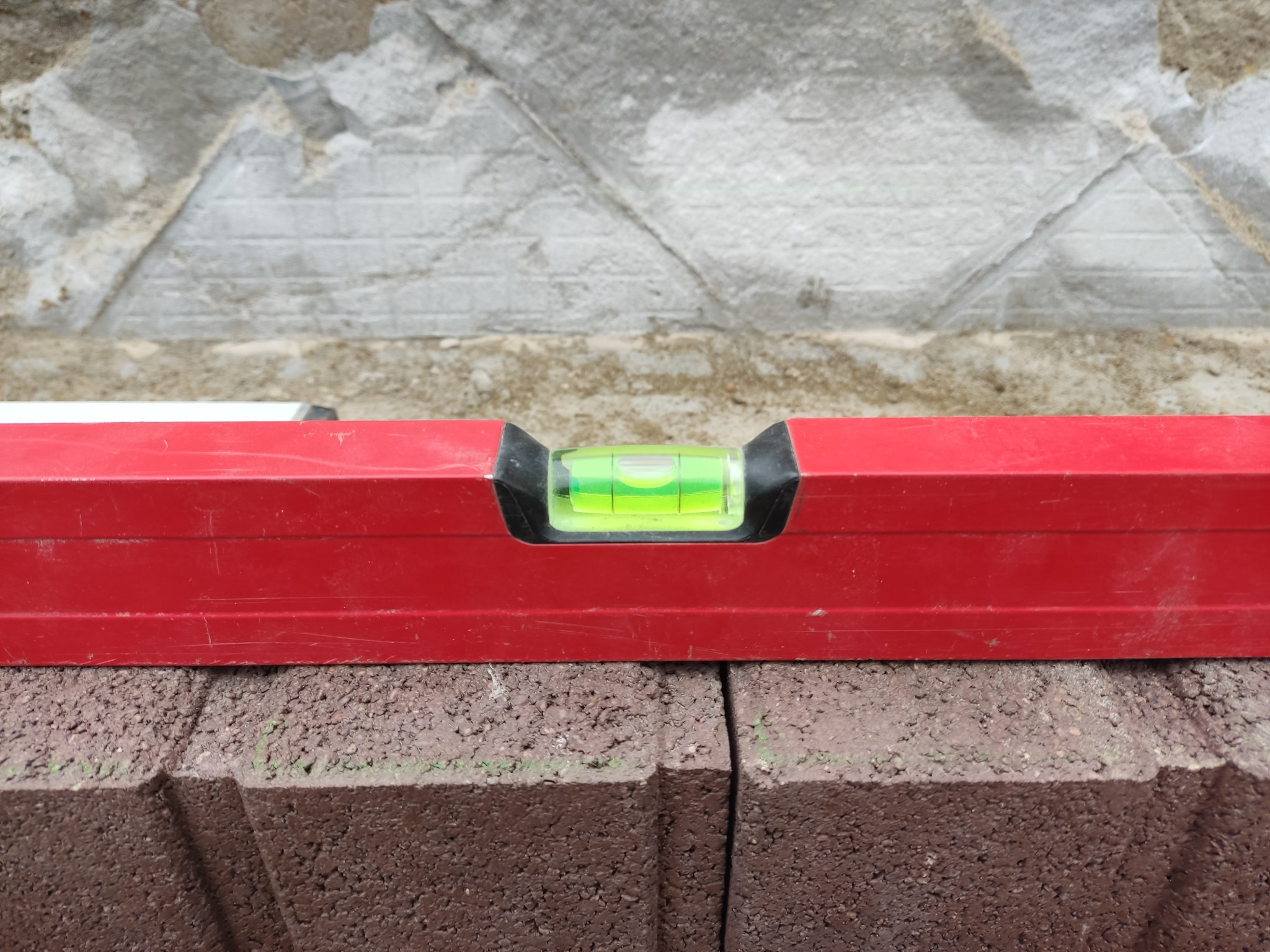 Czerwona poziomica spoczywająca na świeżo ułożonej, brązowej kostce brukowej z widoczną fugą, na tle szarego, surowego muru.