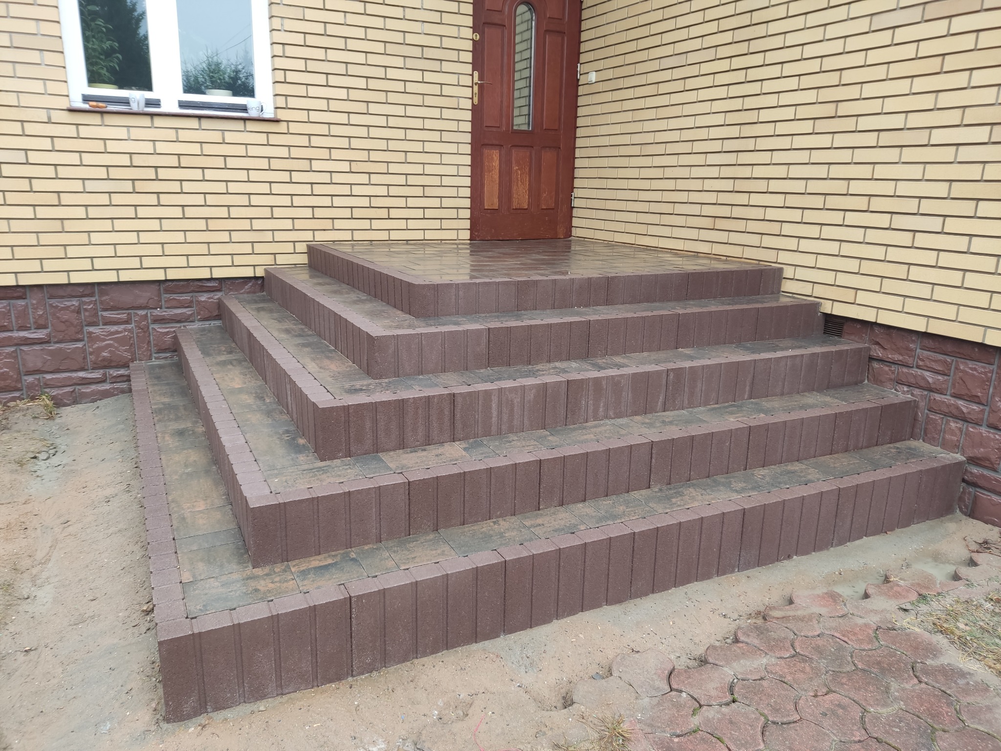 Nowe, czterostopniowe schody wejściowe z brązowej kostki przed budynkiem z żółtej cegły, prowadzące do brązowych drzwi z szybą.