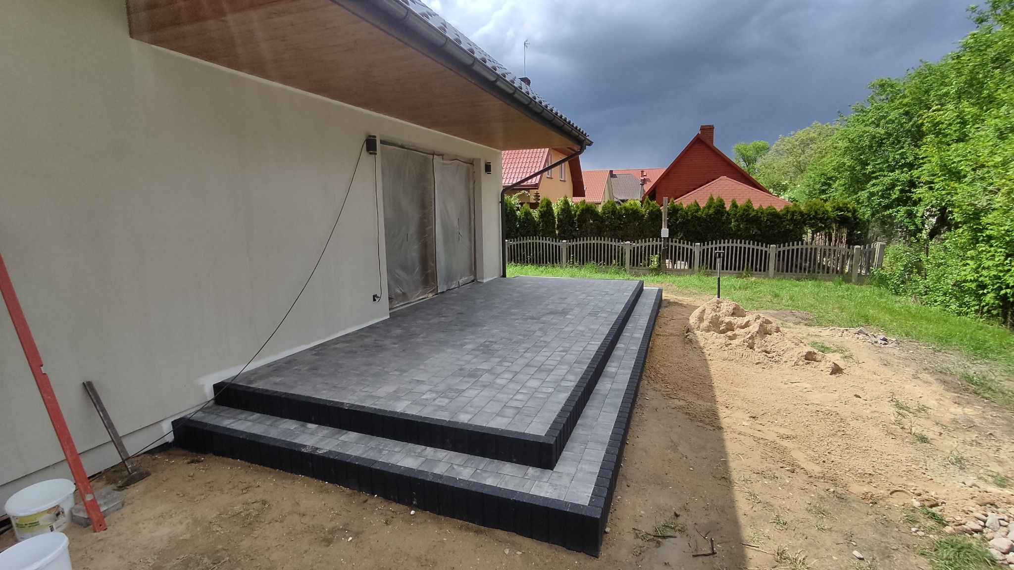 Ułożona kostka brukowa w kolorze szarym tworząca taras z dwoma stopniami przed budynkiem, obrzeża z ciemnej kostki. W tle widoczne budynki mieszkalne i zachmurzone niebo.