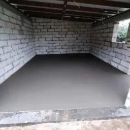 Świeżo wylana, gładka posadzka jastrychowa w surowym pomieszczeniu z białych bloczków, widoczna drewniana konstrukcja dachu i częściowo otwarte okno.