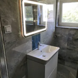 Nowoczesna łazienka z szarą marmurową okładziną, podświetlanym lustrem LED nad białą szafką z umywalką i oknem z widokiem na zieleń