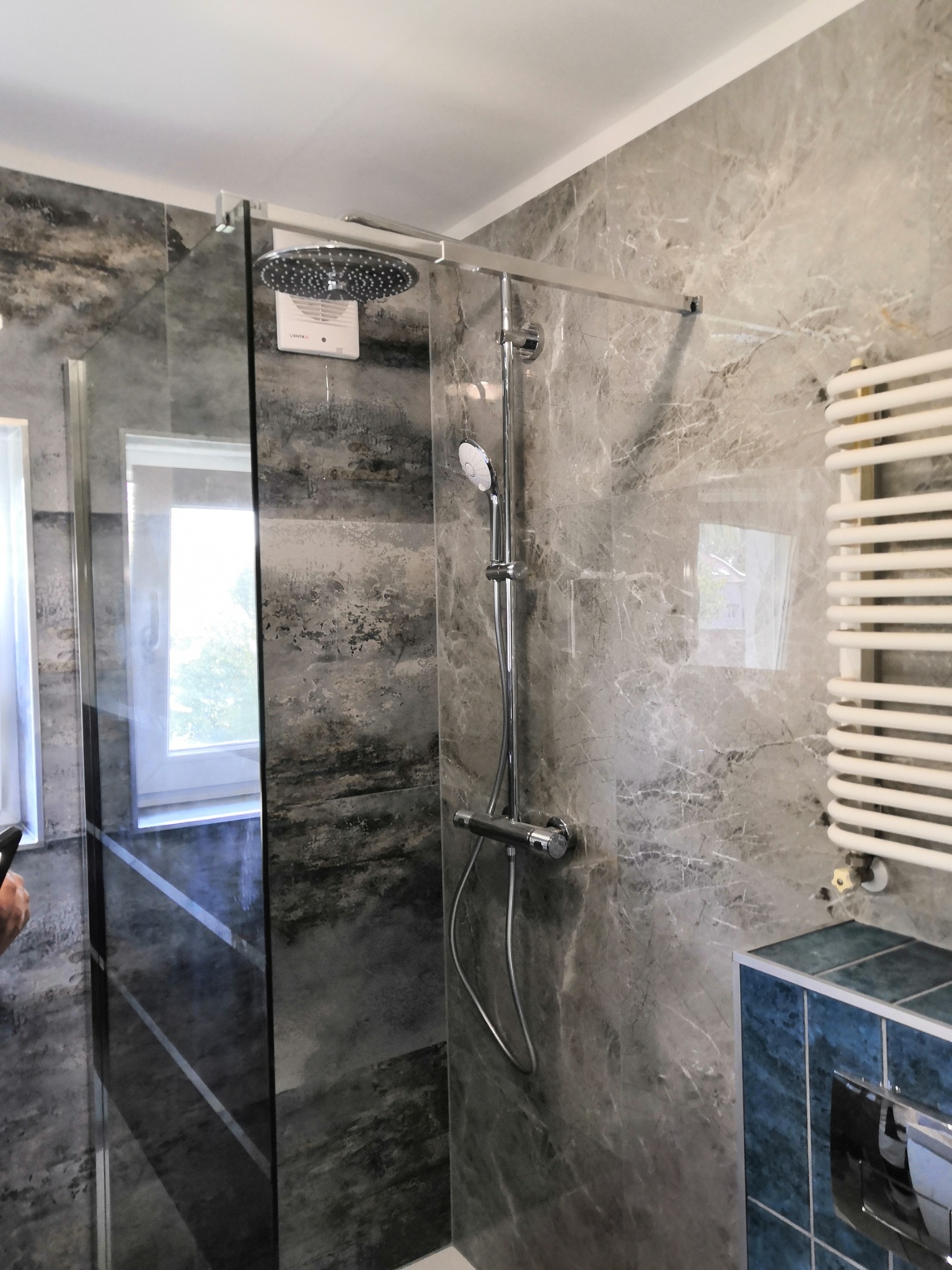 Nowoczesna kabina prysznicowa z deszczownicą i szklanymi drzwiami, wykończona marmurowymi płytkami w odcieniach szarości, z grzejnikiem drabinkowym i zabudowaną spłuczką WC z niebieskimi płytkami.