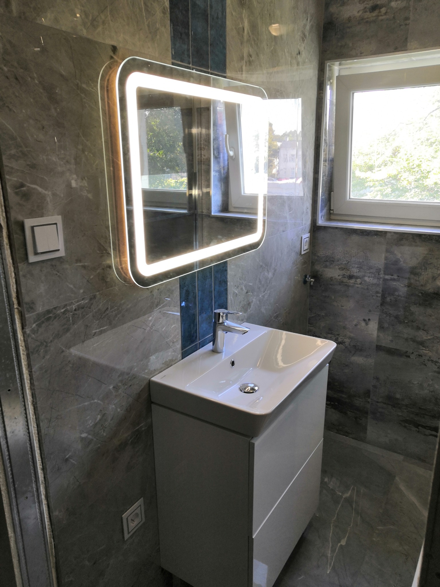 Nowoczesna łazienka z szarą marmurową okładziną, podświetlanym lustrem LED nad białą szafką z umywalką i oknem z widokiem na zieleń