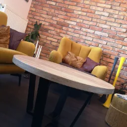 Okrągły stolik kawowy z drewnianym blatem i czarnymi, metalowymi nogami w loftowym salonie z czerwoną ceglaną ścianą i żółtymi fotelami.