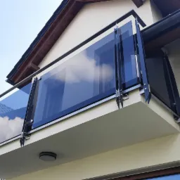 Szklana balustrada balkonowa z czarnymi elementami mocującymi, odbijająca błękitne niebo i chmury, widoczna z dołu na tle jasnej elewacji domu z ciemną dachówką.