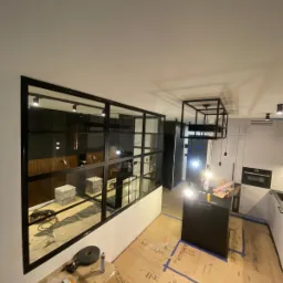 Częściowy widok wnętrza mieszkania w trakcie remontu, z zamontowaną dużą, czarną, przeszkloną konstrukcją działową w stylu industrialnym, oddzielającą przestrzeń, z widocznymi narzędziami...