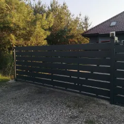 Nowoczesna, pozioma brama wjazdowa w kolorze grafitowym, z widocznym oświetleniem słupków, na tle domu z czerwoną dachówką i zielonych drzew iglastych.