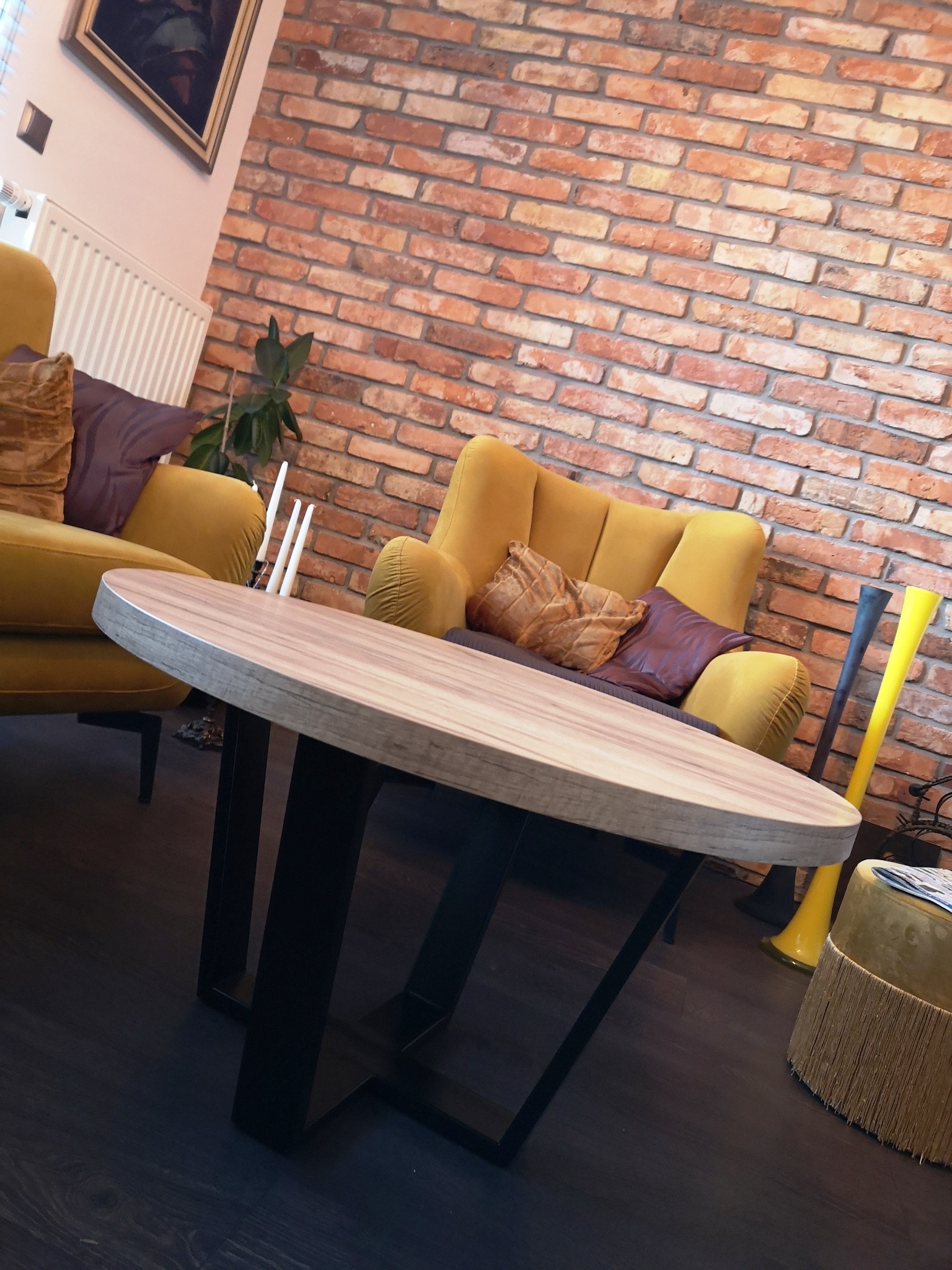 Okrągły stolik kawowy z drewnianym blatem i czarnymi, metalowymi nogami w loftowym salonie z czerwoną ceglaną ścianą i żółtymi fotelami.