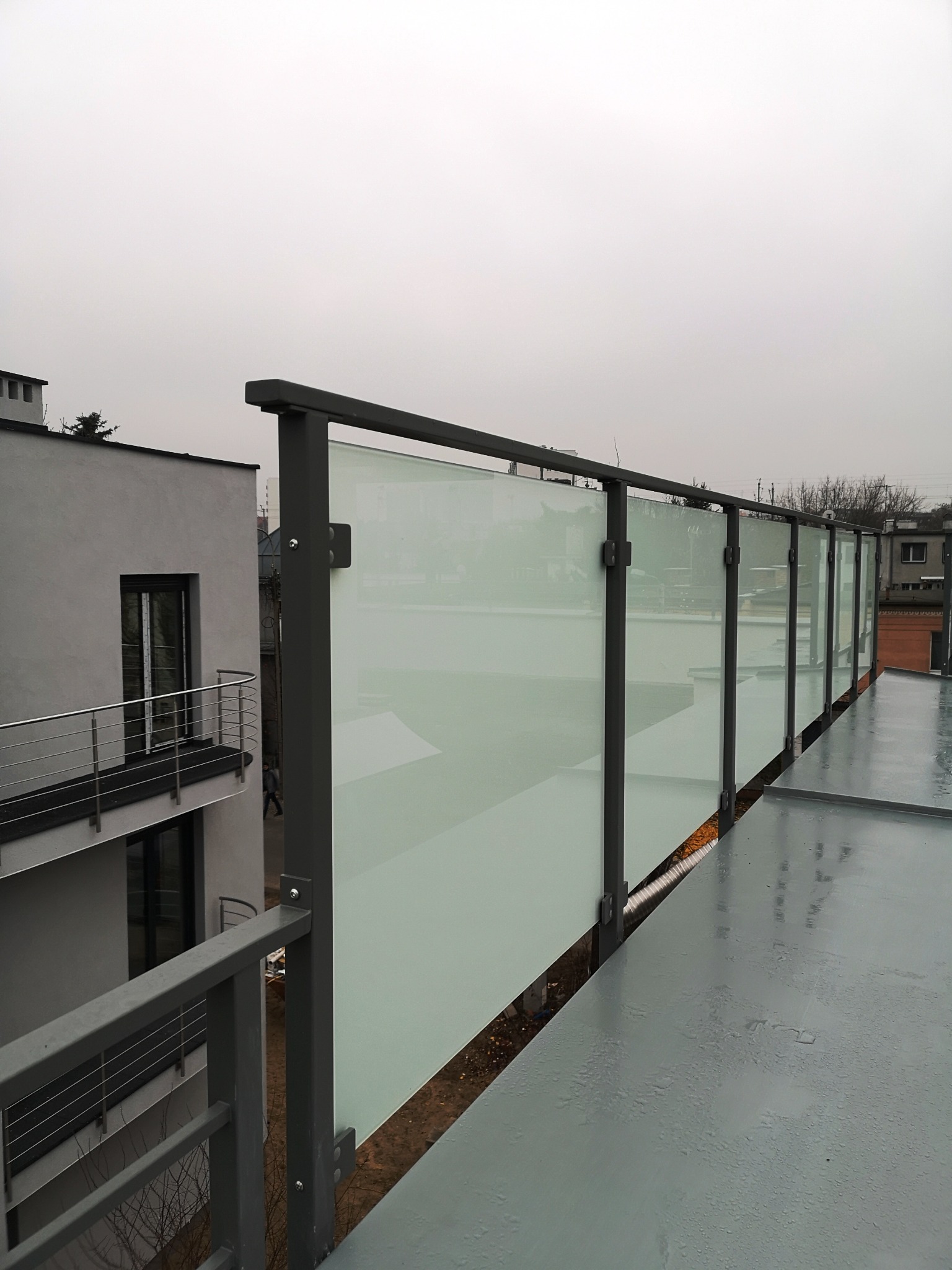 Szklana balustrada z szarymi słupkami na tarasie, widok z góry, pochmurny dzień, w tle budynek z balkonem z metalową balustradą.