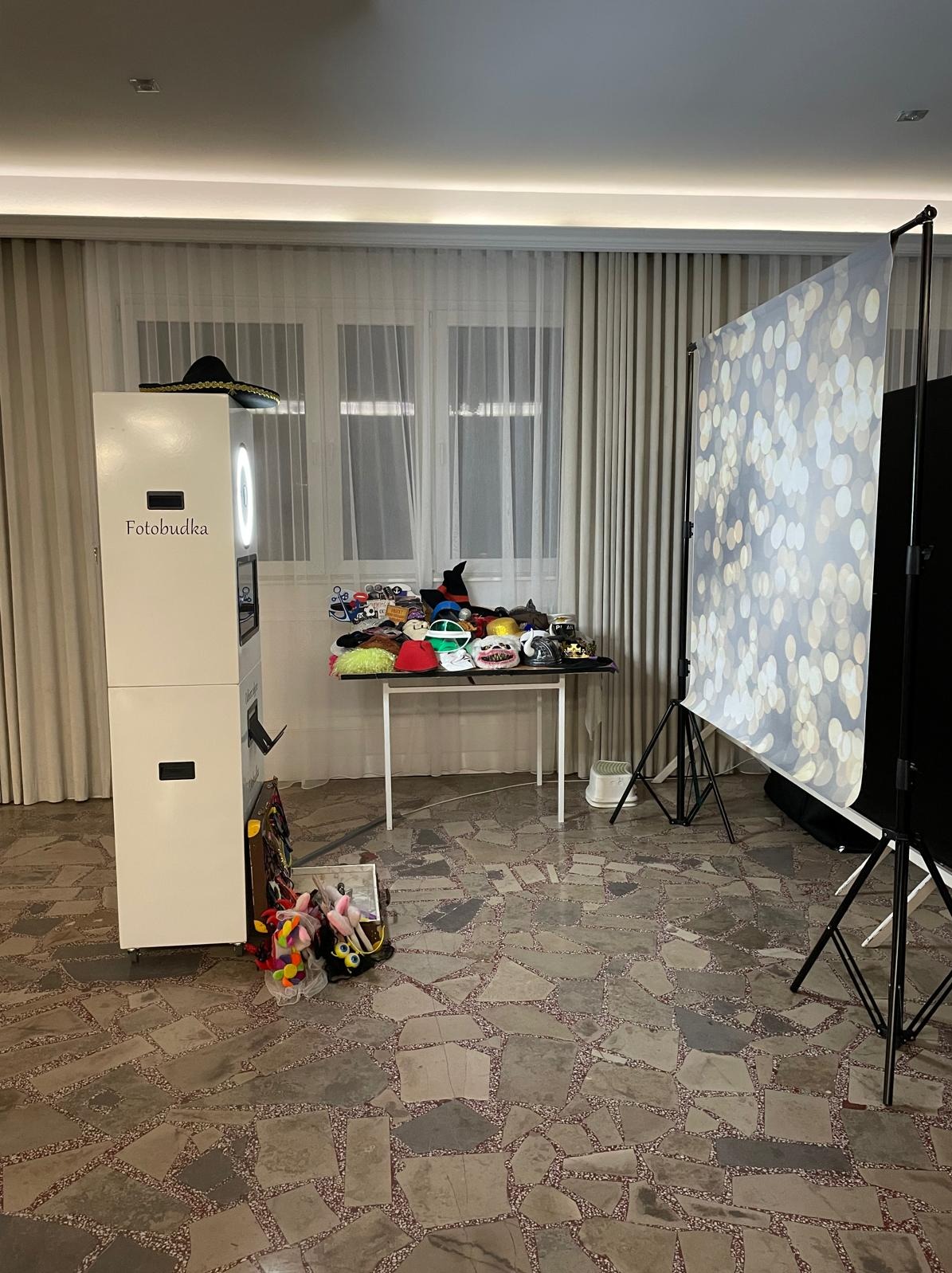 Stanowisko z fotobudką, stolikiem pełnym rekwizytów (kapelusze, maski) i tłem w kropki, gotowe do sesji zdjęciowej na imprezie. Widok z przodu, w pomieszczeniu.