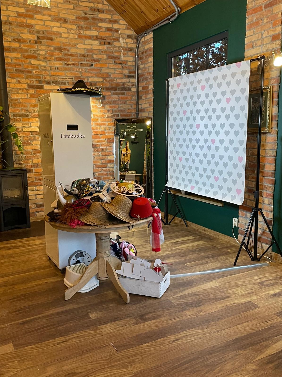 Stanowisko z fotobudką, rekwizytami na okrągłym stole i tłem w serduszka, gotowe do użycia na imprezie w rustykalnym wnętrzu z ceglaną ścianą.