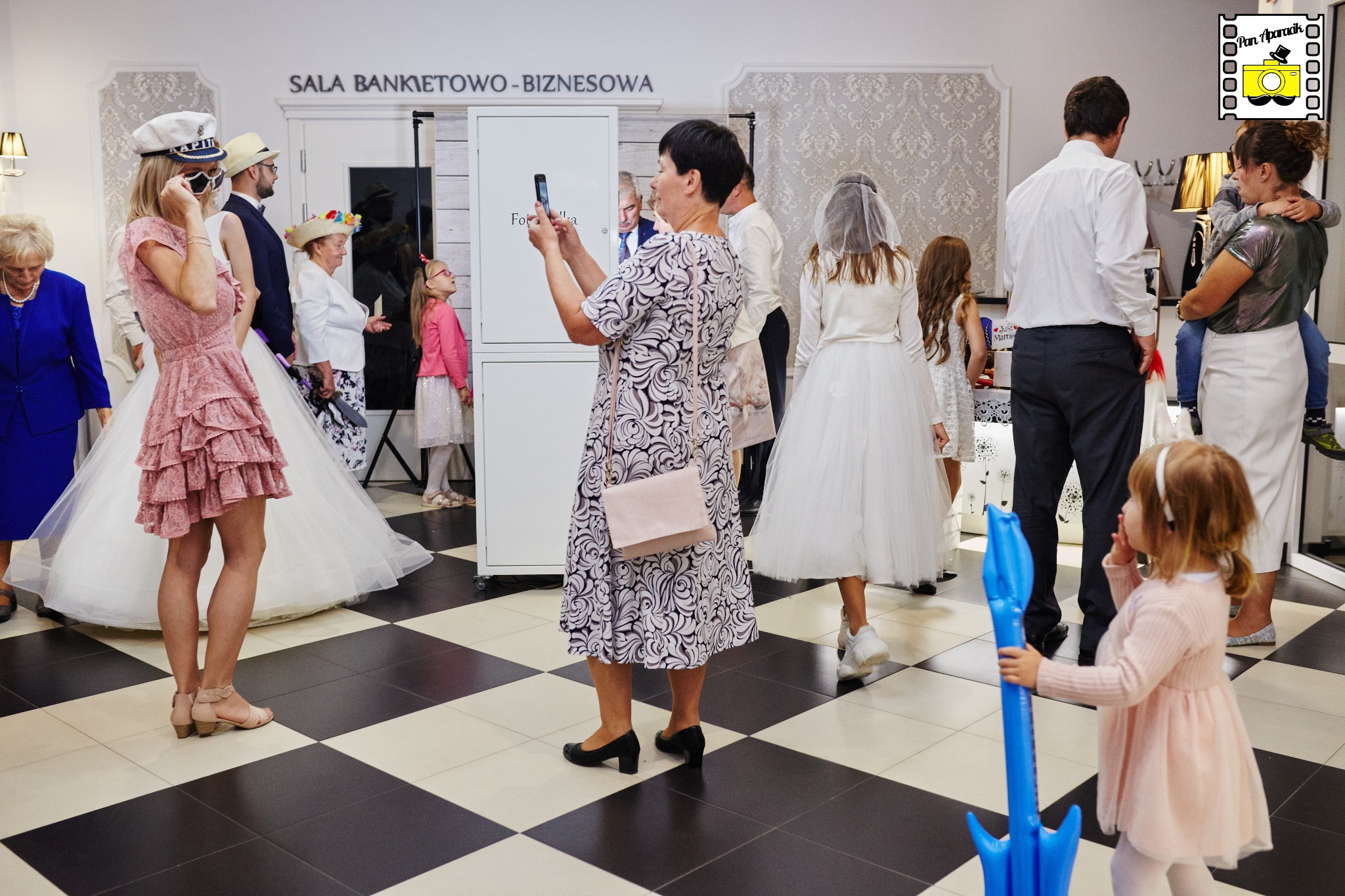 Goście weselni pozują do zdjęć w fotobudce w sali bankietowej. Kobieta robi zdjęcie telefonem. W tle widoczna para młoda i dzieci w strojach komunijnych.