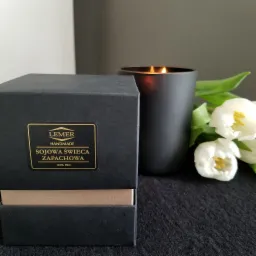 Czarny, otwarty kartonik z etykietą 'LEMER Handmade Sojowa Świeca Zapachowa 100% EKO', obok zapalona, czarna świeca w szkle i białe tulipany na ciemnym tle.