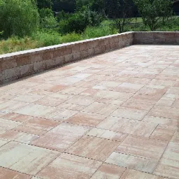 Wyłożony kostką taras z niskim murem oporowym, widok z góry. Na tarasie stoi kolumna i ławka. Widok na zieleń w tle.