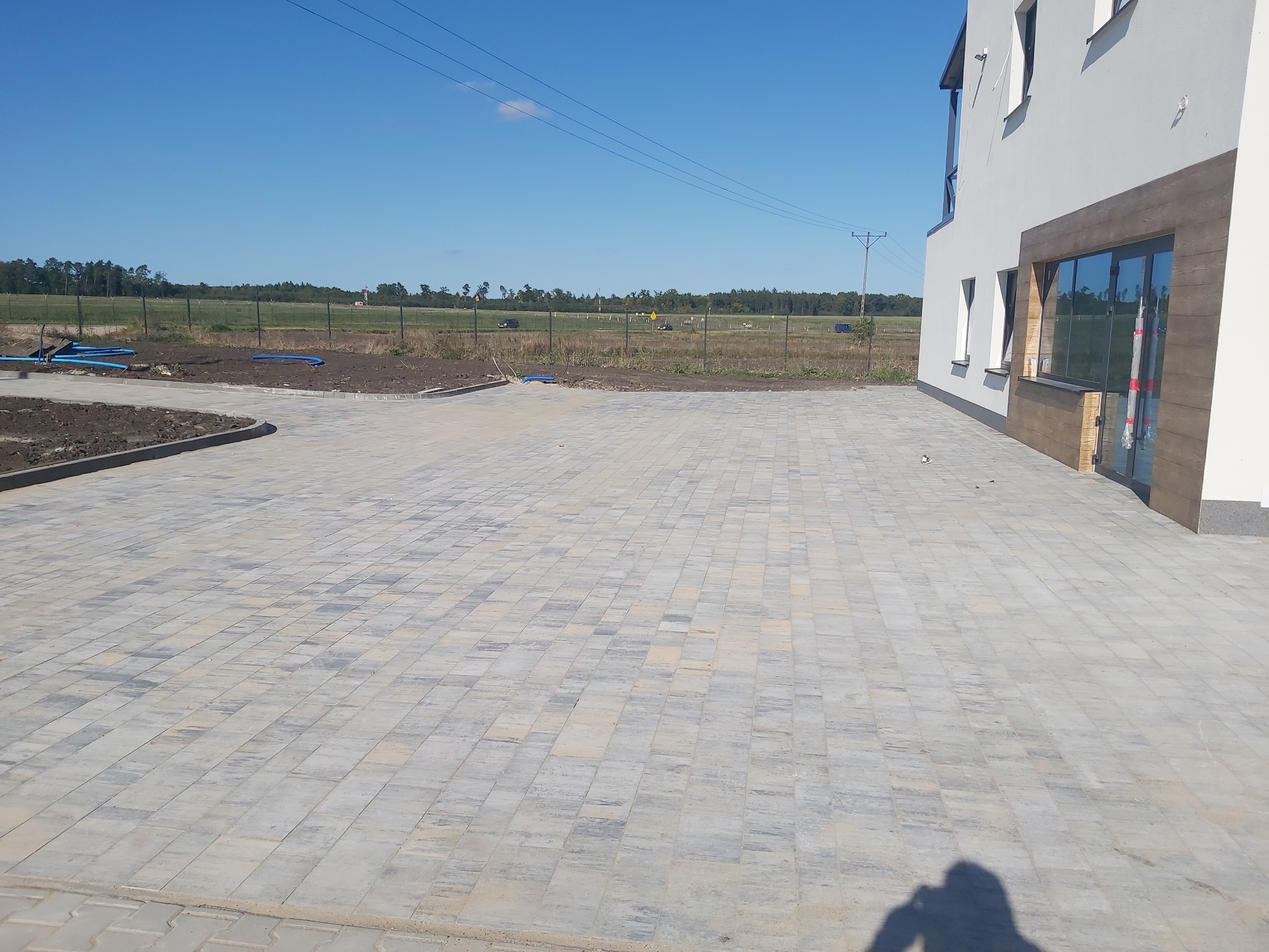 Ułożona kostka brukowa na posesji przy budynku, widoczne krawężniki i przygotowany teren pod dalsze prace. Na horyzoncie drzewa i słupy energetyczne.