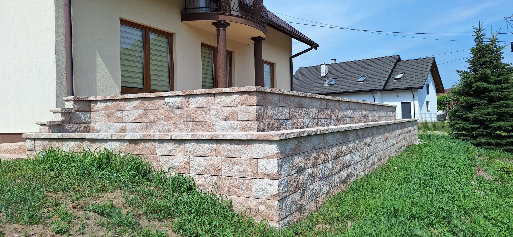 Wykończona obudowa tarasu i schodów wejściowych z kostki łupanej w odcieniach beżu i brązu, na tle domu z balkonem i zieleni. Estetyczne i solidne wykonanie.