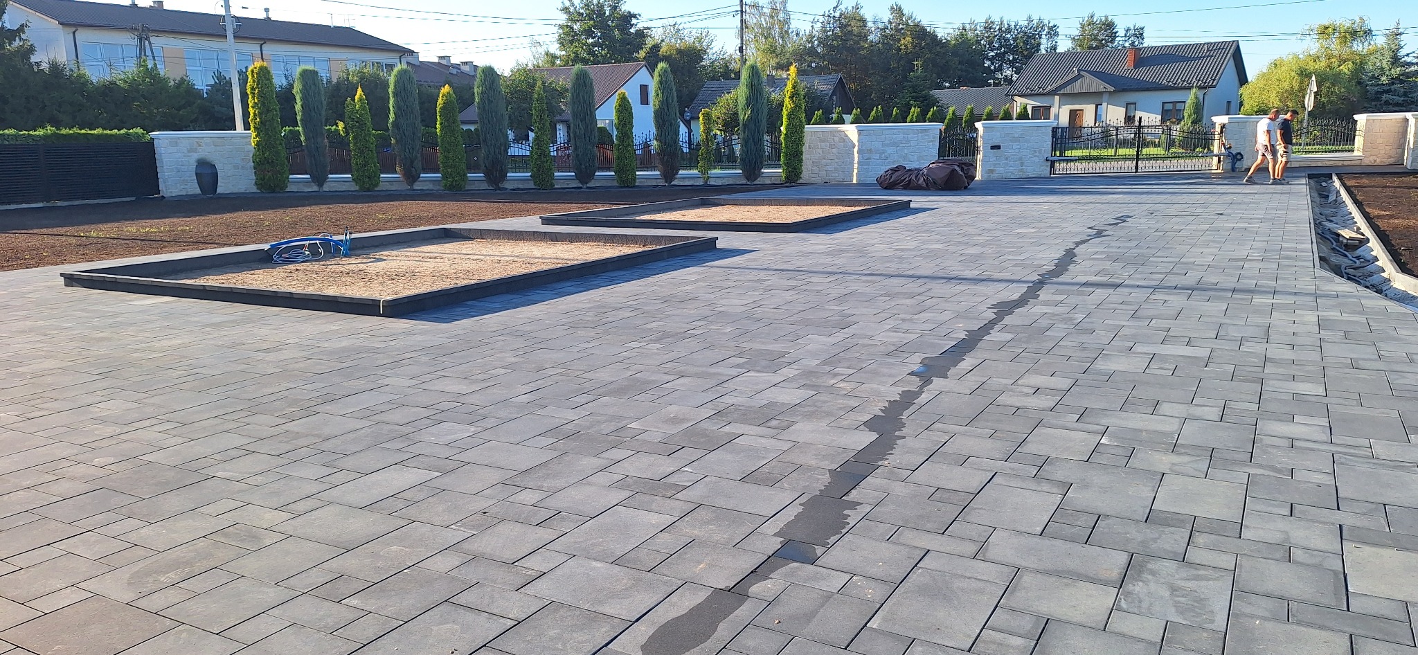 Ułożona, szara kostka brukowa na podjeździe z wyznaczonymi miejscami na zieleń, w tle domy i ogrodzenie z tujami, widoczni ludzie.
