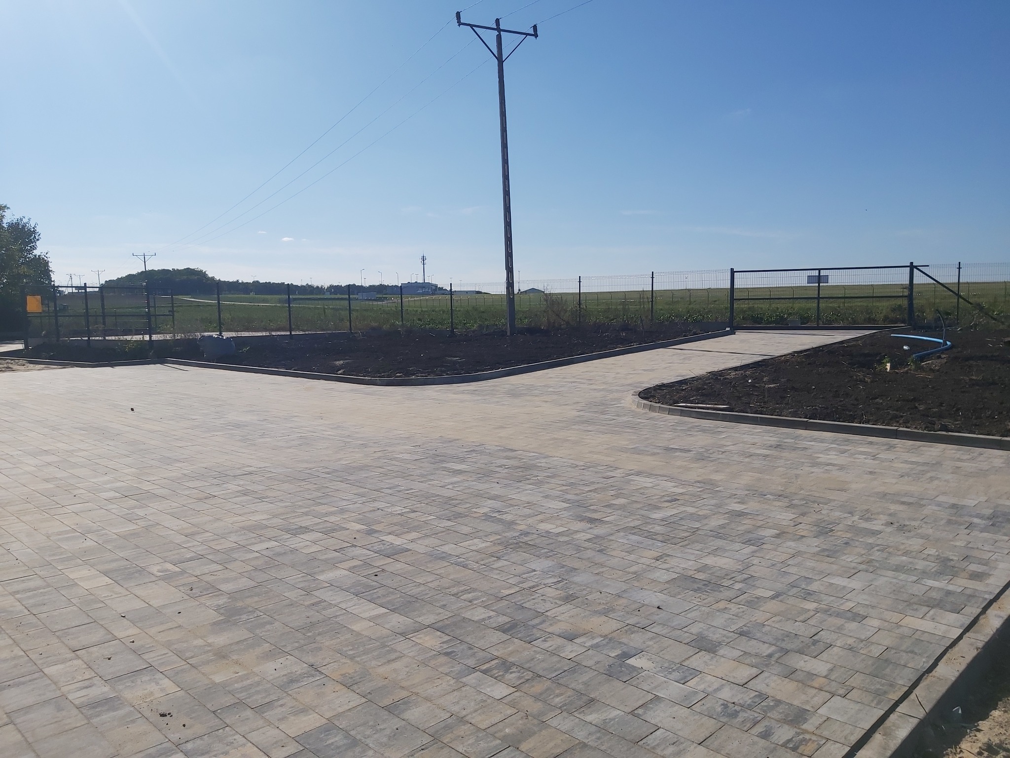 Nowa, szara kostka brukowa ułożona na dużej powierzchni, z obrzeżami i przygotowanym miejscem pod trawnik. W tle ogrodzenie panelowe i słup wysokiego napięcia.