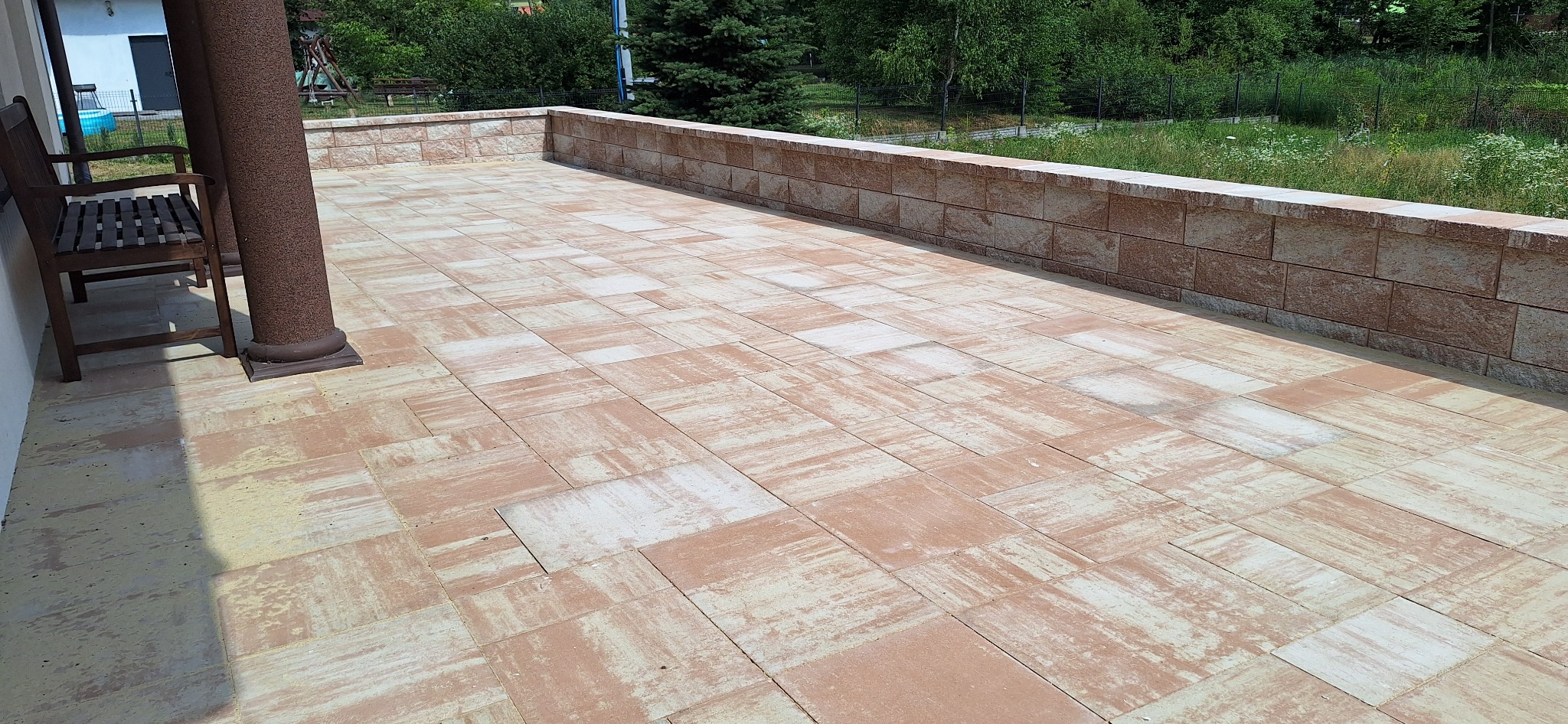 Wykończony taras z kostki brukowej w odcieniach beżu i brązu, otoczony niskim murem z kamienia. Widoczne kolumny podtrzymujące zadaszenie i drewniane krzesło.