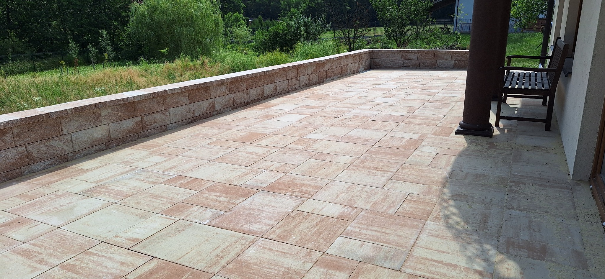Wyłożony kostką taras z niskim murem oporowym, widok z góry. Na tarasie stoi kolumna i ławka. Widok na zieleń w tle.