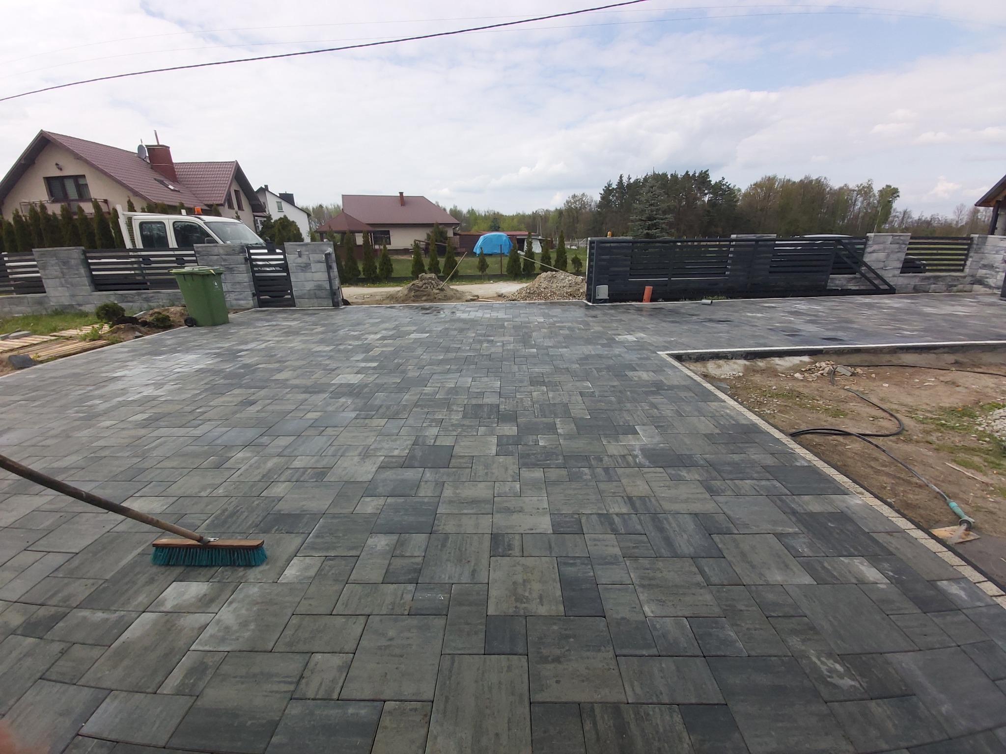 Ułożona kostka brukowa w odcieniach szarości na podjeździe, z widocznym fragmentem szczotki. W tle dom, ogrodzenie z elementami betonowymi i drewnianymi.