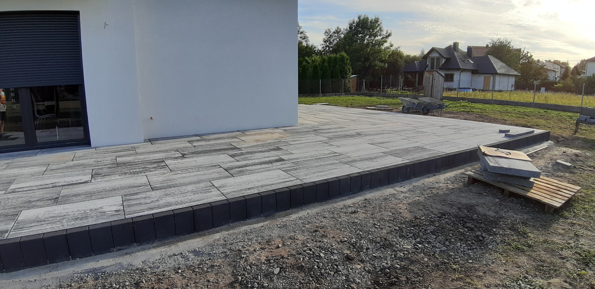 Nowo ułożony taras z szarej kostki brukowej o nieregularnym wzorze, obrzeża z ciemnej kostki, widok na budynek z roletami i ogród z dwoma taczkami w tle.